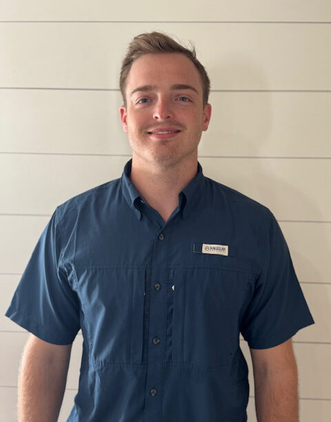A commercial general contractor with short light brown hair, wearing a dark blue short-sleeve button-up shirt, stands in front of a white shiplap wall, looking at the camera and smiling slightly.