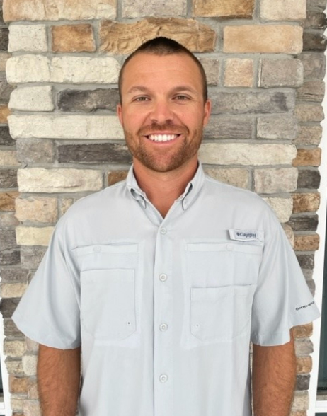 A man with short hair and a trimmed beard stands smiling in front of a stone wall, wearing a light gray button-up shirt with short sleeves, reflecting the approachable professionalism of a commercial general contractor.