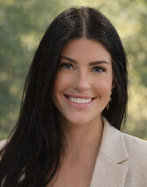 Woman with long dark hair and light eyes, wearing a beige blazer, smiling confidently at the camera with a blurred green background, embodying the professionalism of a commercial general contractor.