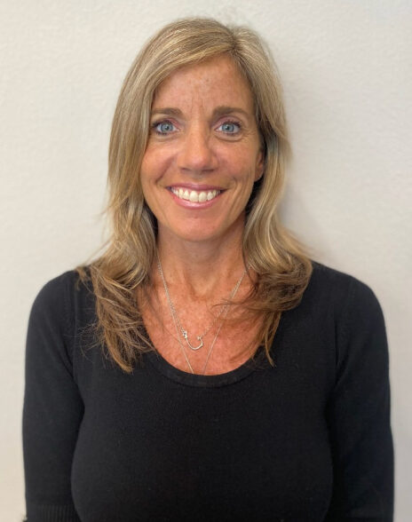 A woman with shoulder-length blonde hair, wearing a black top and silver necklaces, smiles at the camera against a plain light-colored background, reflecting the confidence of a leading commercial general contractor.