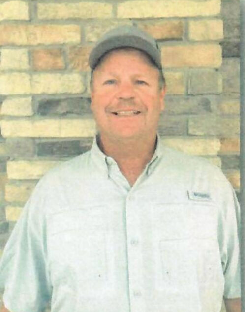 A commercial general contractor wearing a light gray shirt and a gray cap stands smiling in front of a brick wall.